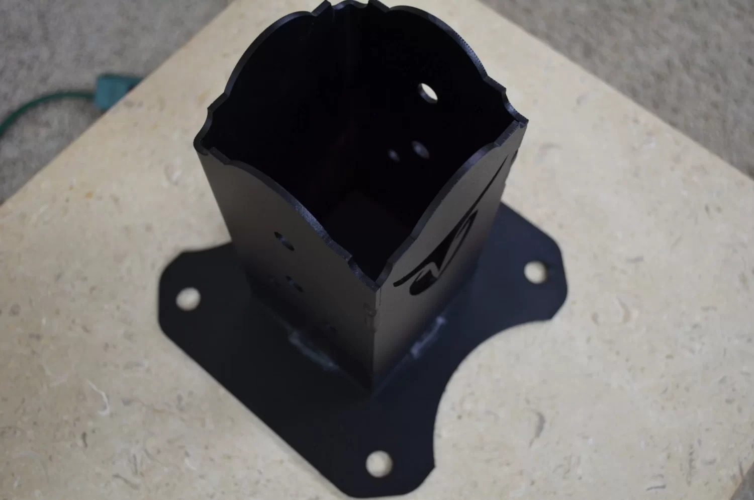 The Universal 4x4 Post Anchor Bracket, featuring a black finish, square hollow center, rounded edges, and four mounting holes, is shown on a beige textured surface.