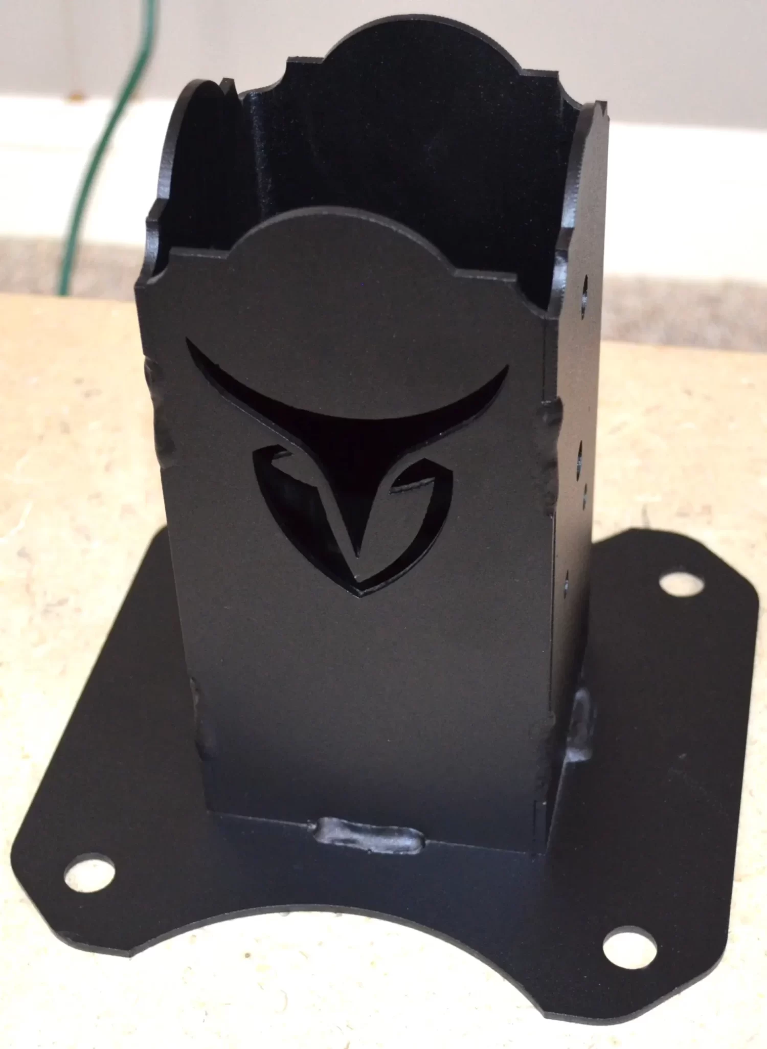 The Universal 4x4 Post Anchor Bracket, featuring a black metal finish and a stylized cutout logo on its front, is shown mounted on a square base plate with four corner holes and displayed on a beige surface.