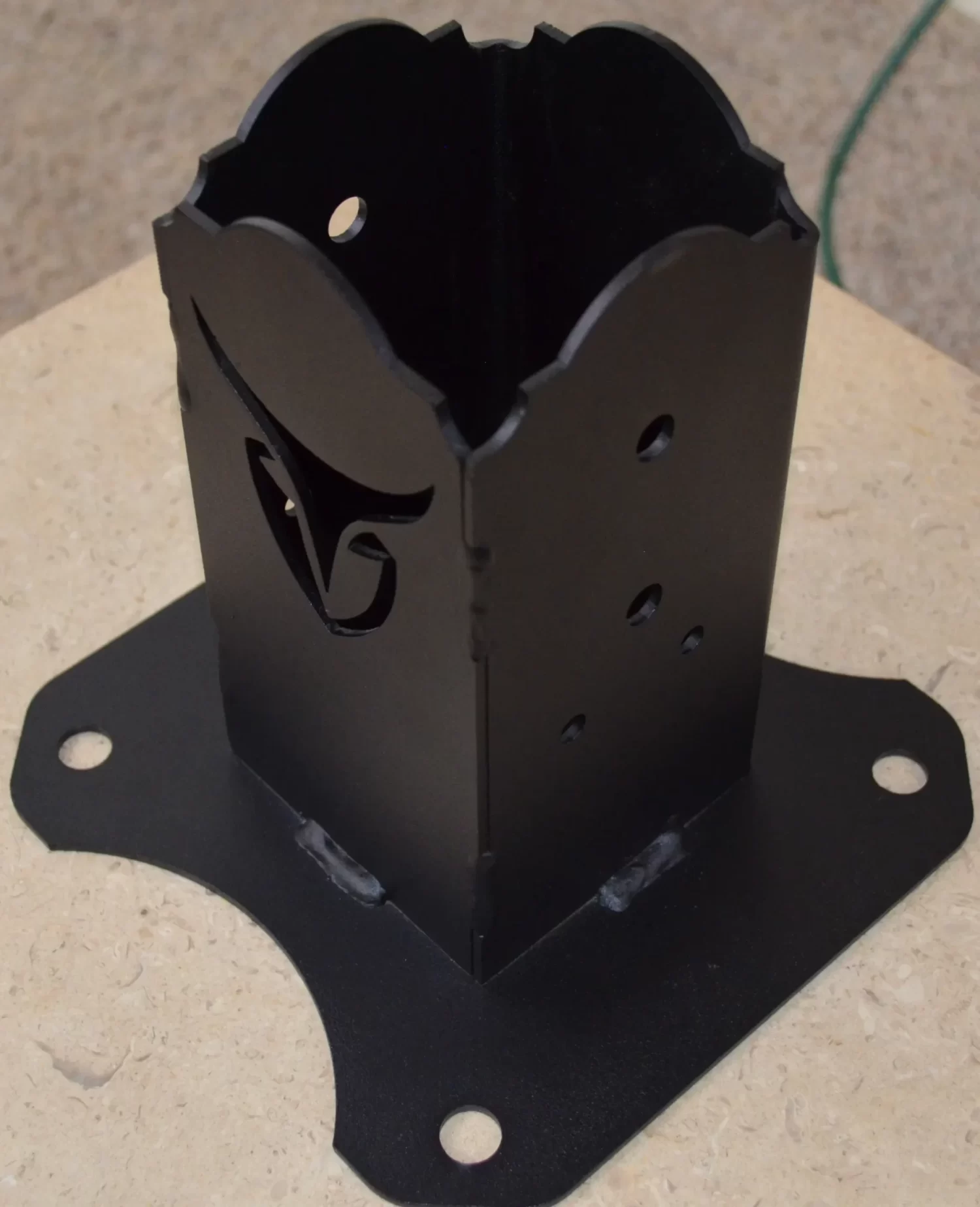 The Universal 4x4 Post Anchor Bracket is a black metal bracket with decorative cutouts, a flared base, and four mounting holes at each corner, shown mounted on a square beige surface for secure installation.