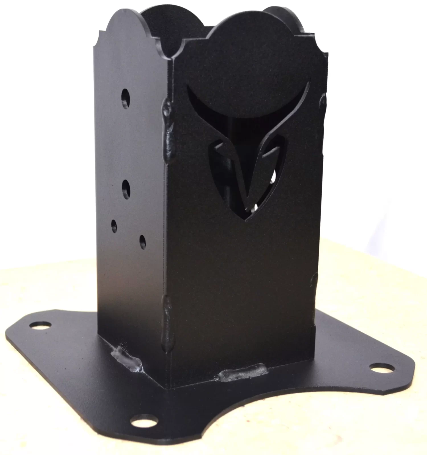 The Universal 4x4 Post Anchor Bracket is a black metal bracket with a wide base, multiple holes, and an owl face cutout design on one side, shown resting on a light-colored surface.