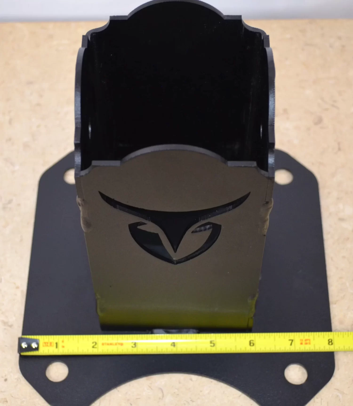 The Universal 4x4 Post Anchor Bracket is black metal with a stylized logo cutout, has a flat base with four bolt holes, and measures about 8 inches wide. A yellow measuring tape is shown at the base on a tan surface.