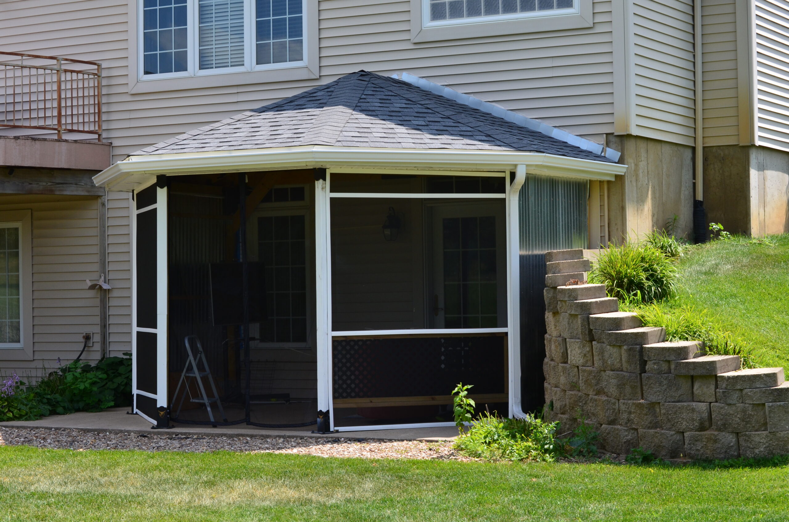 DSC_0026 A Heavy Duty 4x4 45-Degree Octagon Elbow Bracket supports a wall-mounted partial octagon screened patio with a sloped roof attached to the back of a beige house, bordered by a small retaining wall, greenery, and a neat lawn.