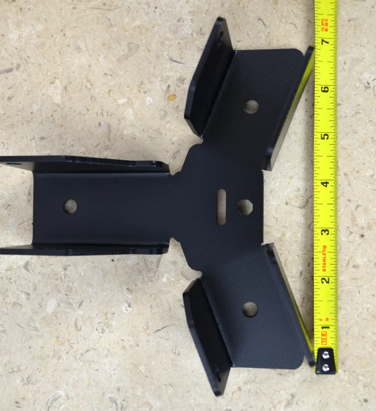 A Heavy Duty Octagon CrossTie Bracket in black metal with multiple holes is placed on a beige textured surface next to a yellow measuring tape showing about 6 inches.