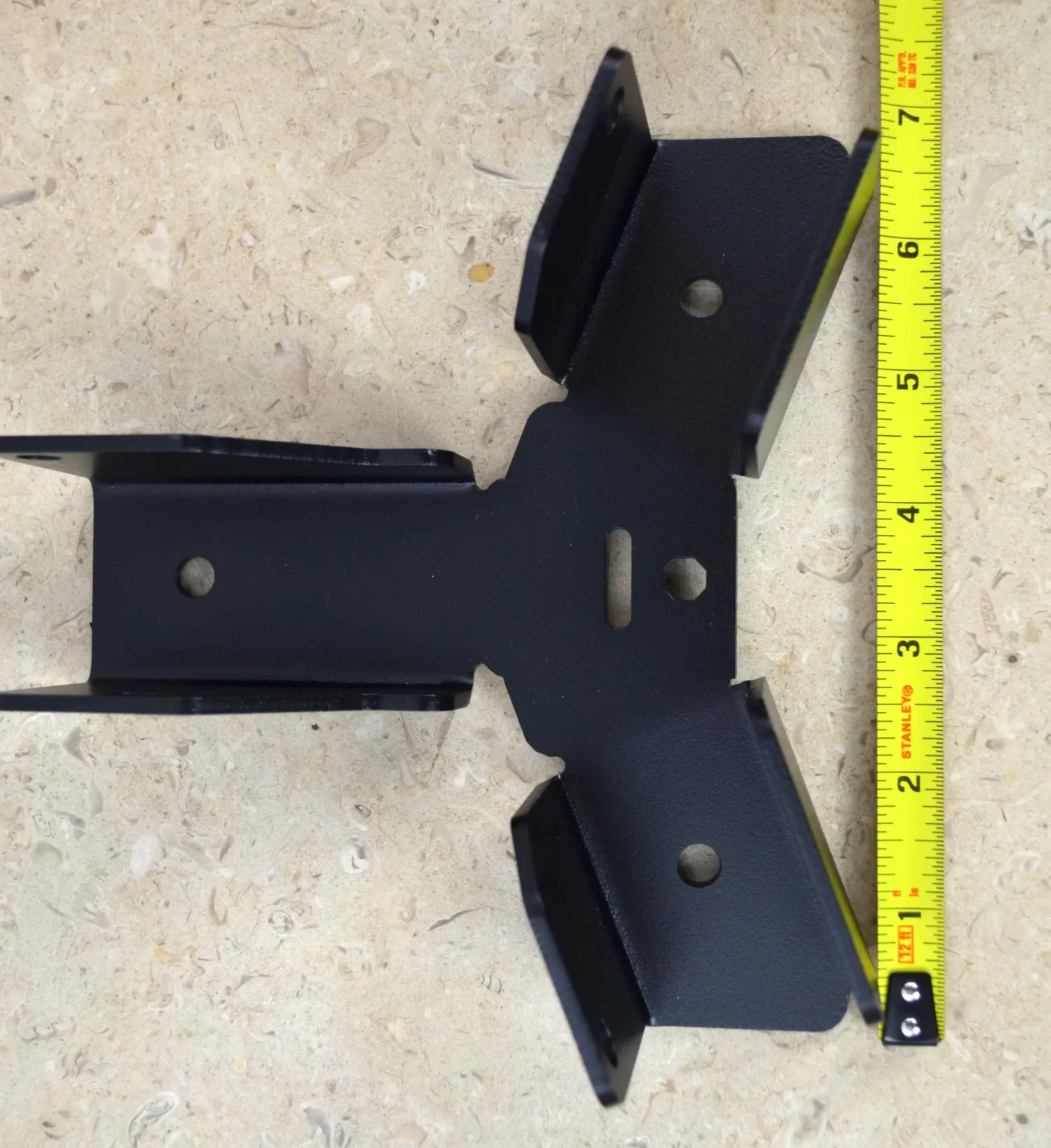 DSC_0092 A Heavy Duty Octagon CrossTie Bracket in black metal with multiple holes is placed on a beige textured surface next to a yellow measuring tape showing about 6 inches.