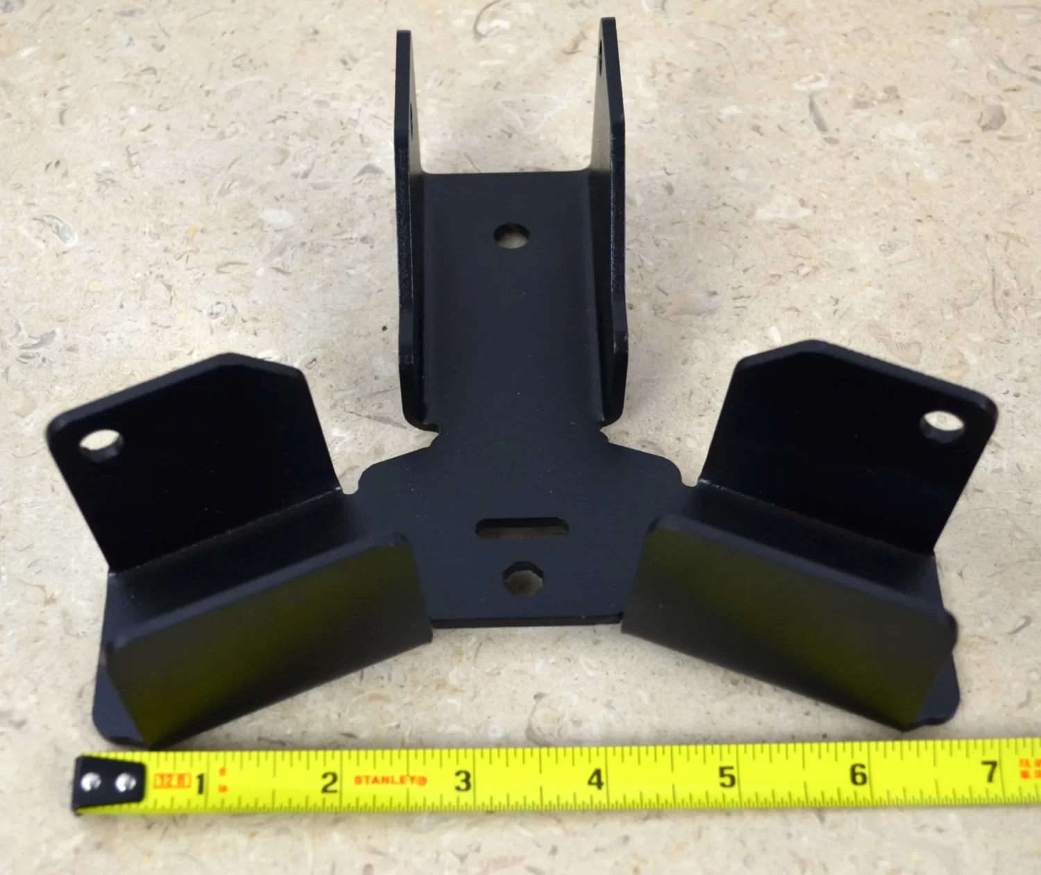 DSC_0095 A Heavy Duty Octagon CrossTie Bracket in black, featuring three angled sections, sits on a beige textured surface next to a yellow measuring tape showing its width is about 8 inches.
