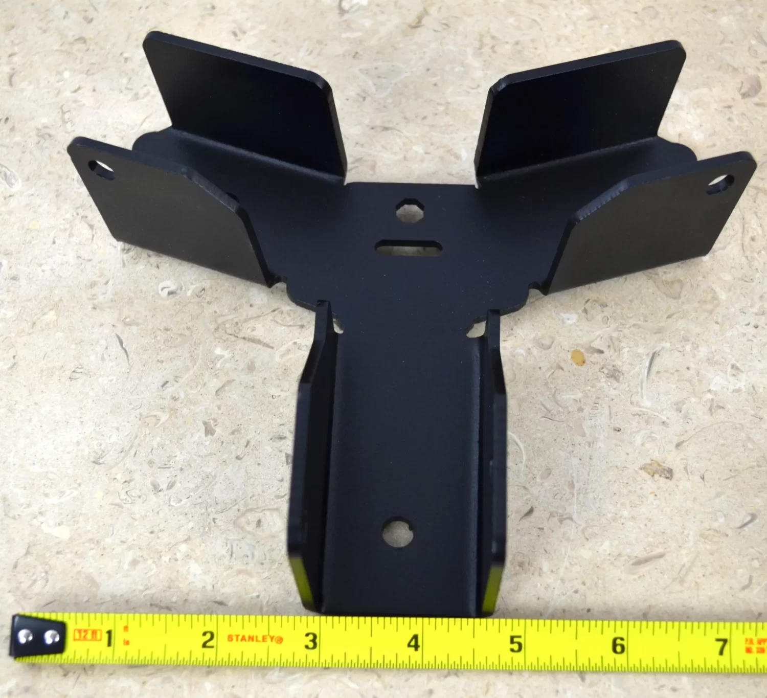 DSC_0098 A Heavy Duty Octagon CrossTie Bracket with three flanges rests on a beige speckled surface. A yellow measuring tape beneath the black metal bracket shows its width is approximately 7 inches.