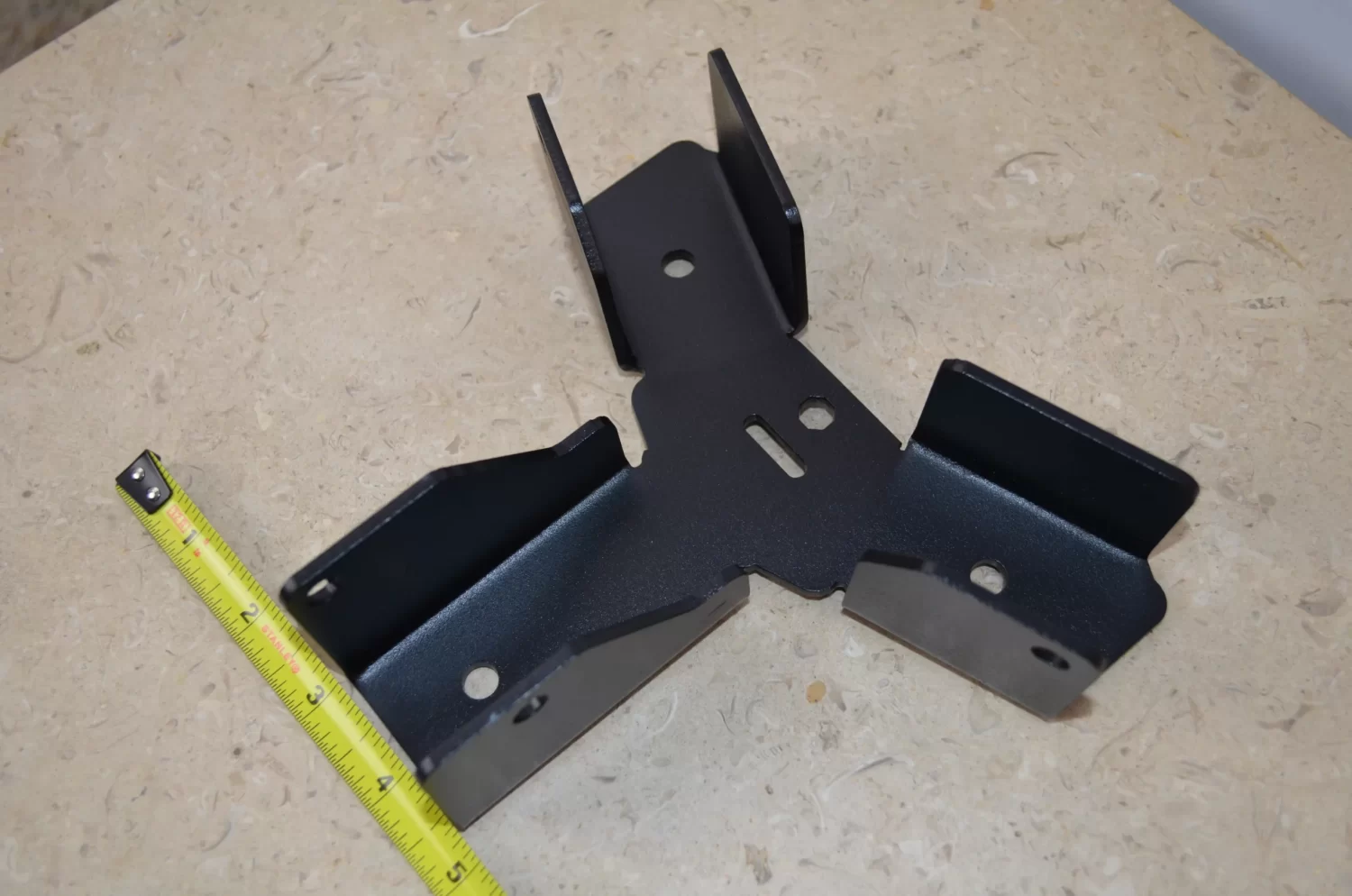 DSC_0103 A Heavy Duty Octagon CrossTie Bracket with three angled arms and multiple holes rests on a beige textured surface, with a yellow measuring tape beside it for scale.