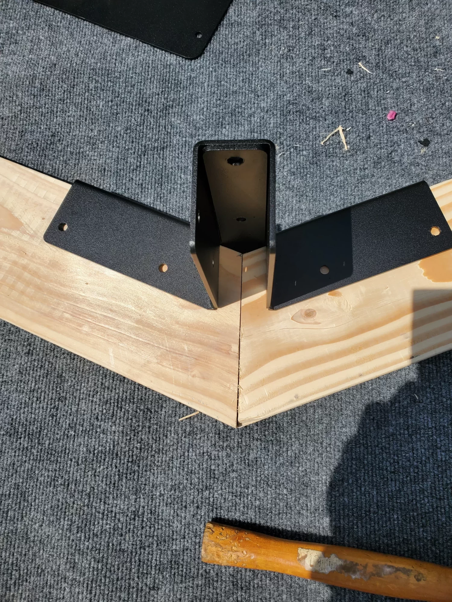 20230510_120314 A Heavy Duty Gable-End Truss Bracket for 4-12 Pitch Rafters is attached at the joint of two wooden boards forming a right angle, resting on gray carpet. A wooden-handled tool appears partially in the lower right corner.