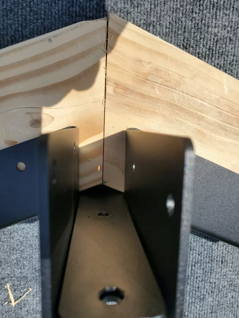 Close-up of two pieces of unfinished wood joined at a right angle with a Heavy Duty Gable-End Truss Bracket for 4-12 Pitch Rafters positioned underneath, showing pre-drilled holes for mounting on a grey textured background.