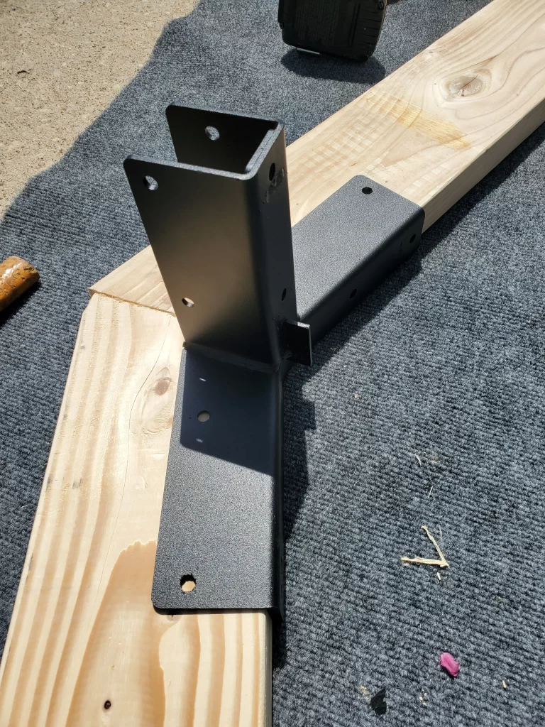 A Heavy Duty Gable-End Truss Bracket for 4-12 Pitch Rafters is attached to two wooden beams in an L-shape, lying on a dark carpet with debris and tools nearby under bright sunlight—ideal for your next DIY project.