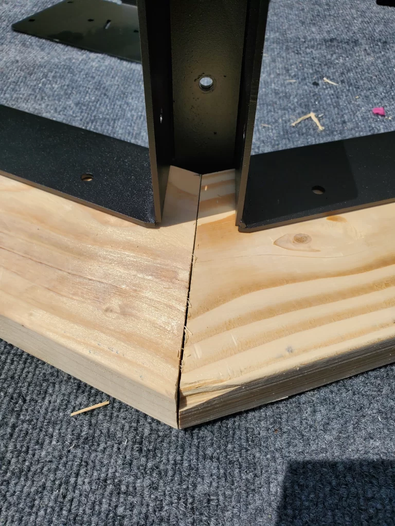 Two pieces of light wood joined at a 45-degree angle feature a Heavy Duty Gable-End Truss Bracket for 4-12 Pitch Rafters attached on top, placed on gray carpet. Some wood shavings are nearby.