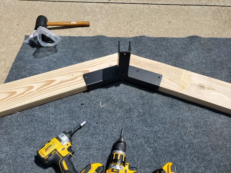 Two wooden beams are joined at an angle using the Heavy Duty Gable-End Truss Bracket for 4-12 Pitch Rafters. Two yellow power drills and a black rubber mallet rest nearby on a gray mat atop a concrete surface.