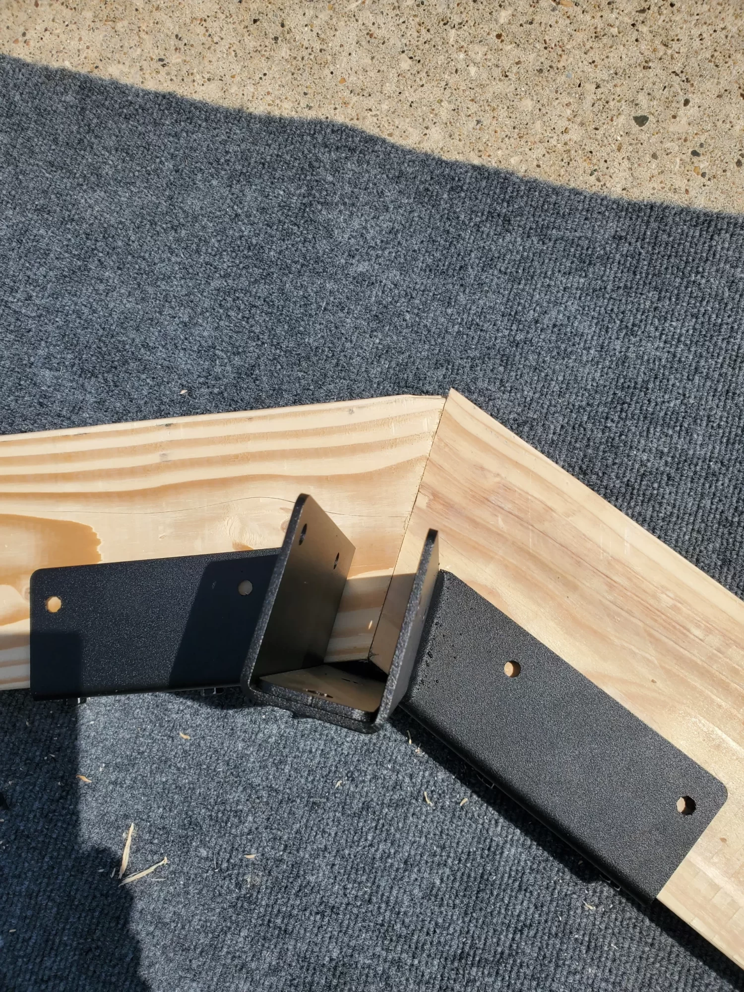 20230510_120715 Two light wood pieces are joined using two Heavy Duty Gable-End Truss Brackets for 4-12 Pitch Rafters, each bracket featuring screw holes. The assembly rests on dark fabric with a concrete surface visible in the background.