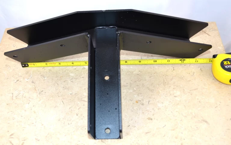 The 5-12 Pitch Gable End Truss Rafter Bracket, made of black metal with three T-shaped arms, is shown on a light wooden surface with a yellow measuring tape behind it to indicate its width.