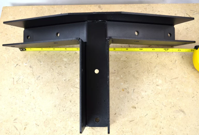 A Heavy Duty Gable-End Truss Bracket for 4-12 Pitch Rafters rests on a beige textured surface. A yellow measuring tape behind the black, multi-holed bracket shows its width. Ideal for DIY projects and secure rafter mounting.