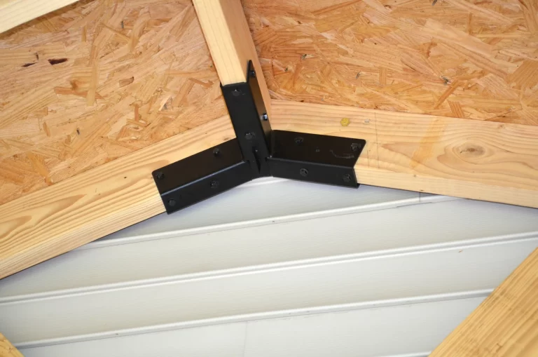 Close-up of wooden roof trusses, made from OSB sheathing and solid lumber, joined at an angle above white horizontal siding using the Heavy Duty Gable-End Truss Bracket for 4-12 Pitch Rafters - DIY Project.