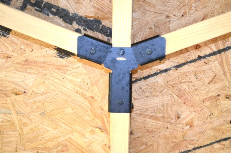 Close-up of a Heavy Duty Octagon CrossTie Bracket joining three light wooden beams on an OSB surface. The bracket is secured with screws, creating a strong triangular connection.