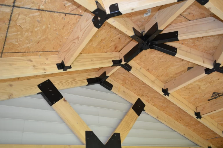 Close-up of a wooden truss roof with Heavy Duty Gable-End Truss Bracket for 4-12 Pitch Rafters connecting beams to a plywood ceiling. A section of gray, horizontally-paneled wall is seen below the structure.