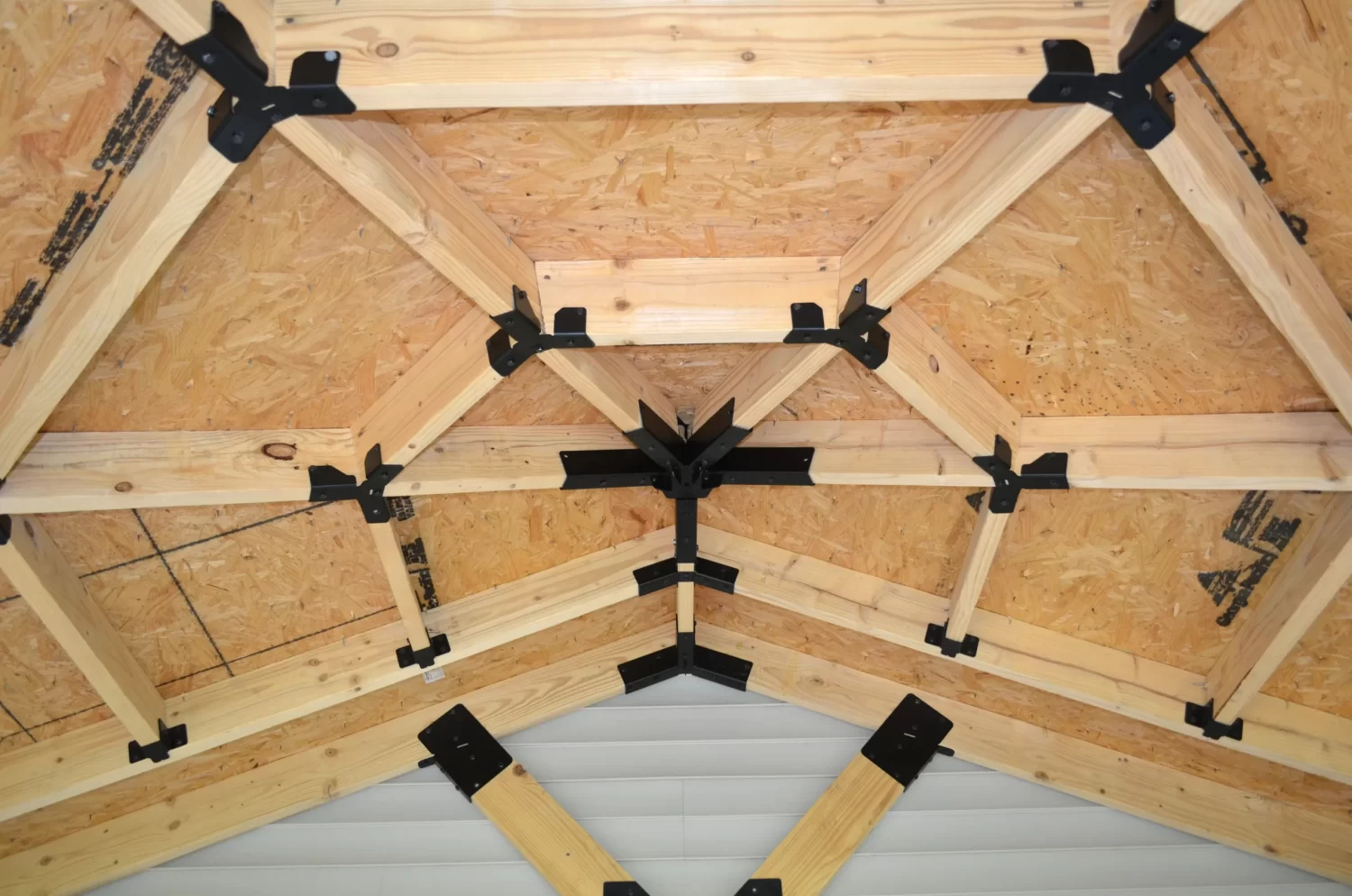DSC_0062 Close-up of a wooden roof truss system featuring light-colored beams and Heavy Duty Octagon CrossTie Brackets at the joints, highlighting the geometric structure and support beneath plywood sheathing.