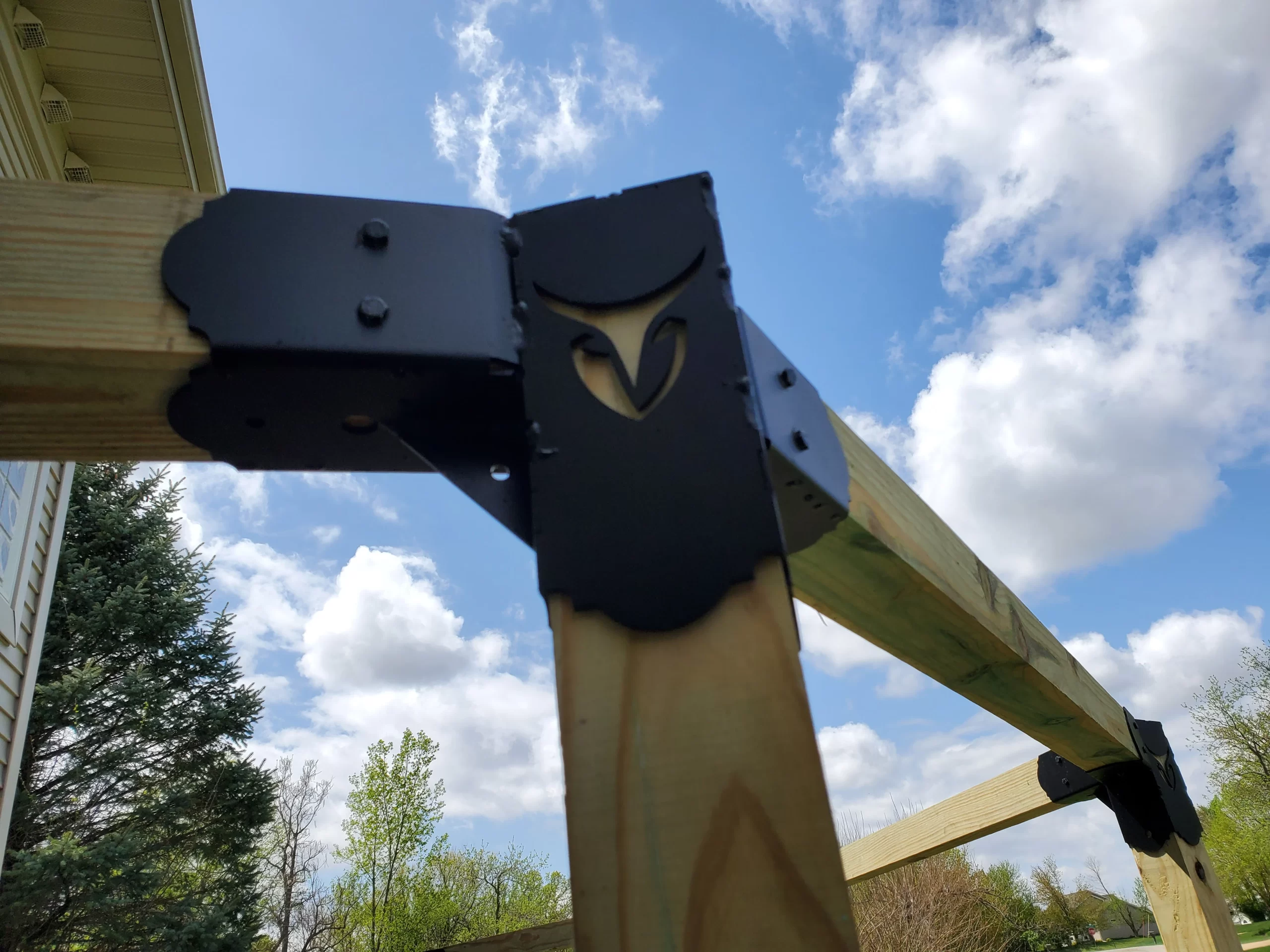 20230508_135713 Close-up of a Heavy Duty 4x4 45-Degree Octagon Elbow Bracket with cutout design, securing two wooden beams outdoors beneath a blue sky with scattered clouds and trees in the background.