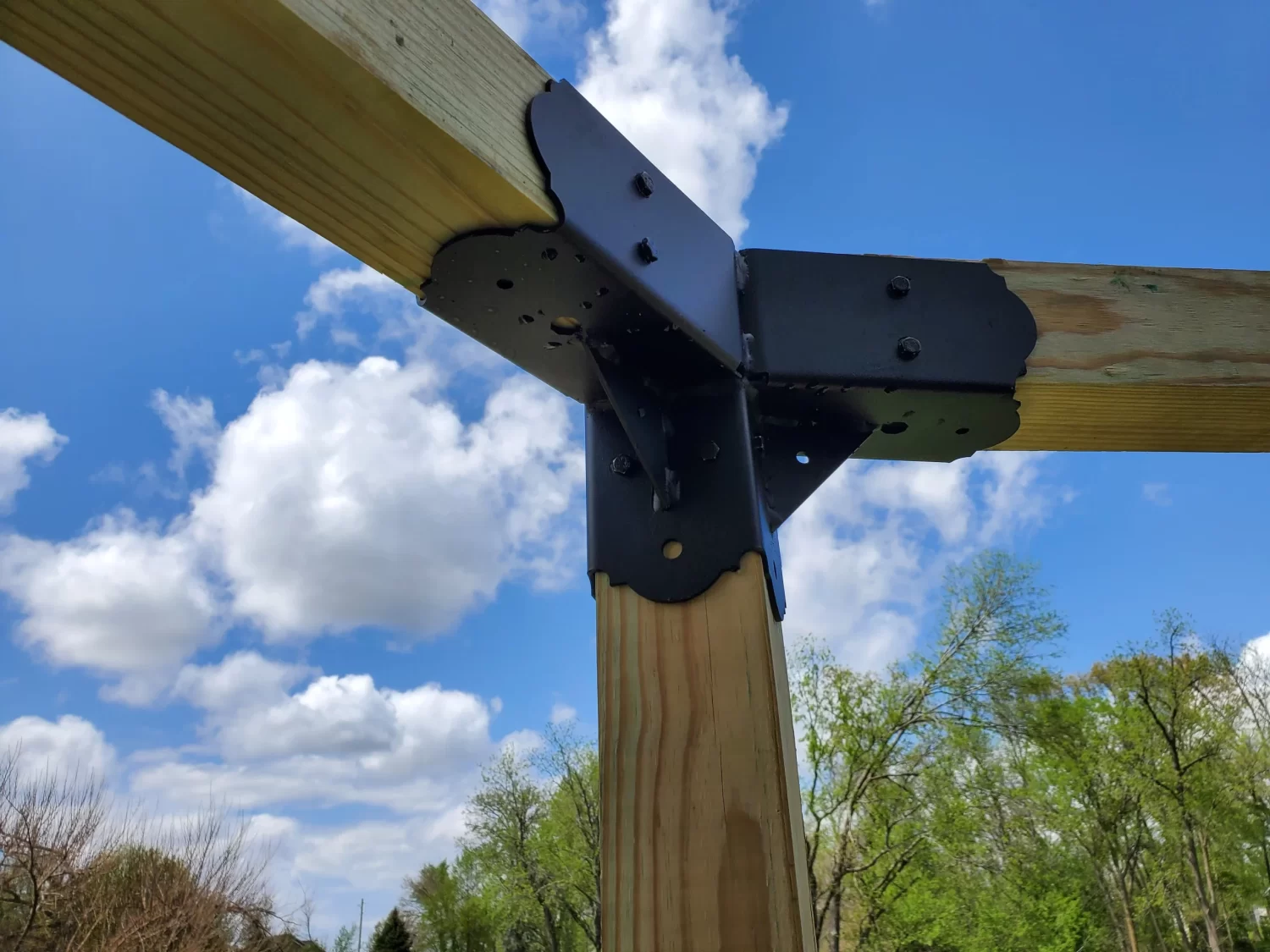 20230508_135749 Close-up of a Heavy Duty 4x4 45-Degree Octagon Elbow Bracket joining three wooden beams at a joint, with a blue sky and clouds above green trees in the background.