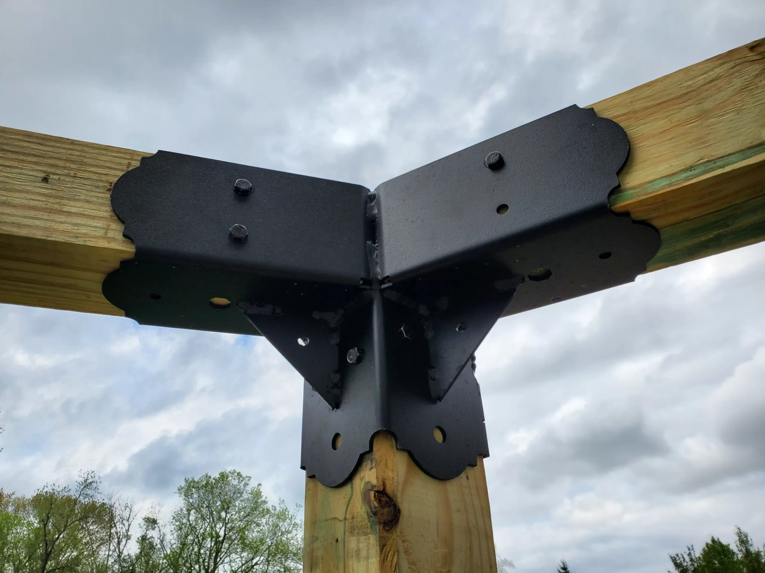 20230508_172421 Close-up of a Heavy Duty 4x4 45-Degree Octagon Elbow Bracket connecting three large wooden beams at a corner joint outdoors, framed by a cloudy sky and trees in the background.