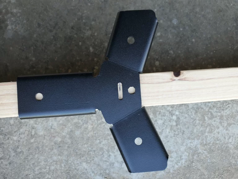 The Heavy Duty Octagon CrossTie Bracket in black connects three wooden beams at right angles on a concrete surface, demonstrating robust construction hardware for framing or building sturdy structures.