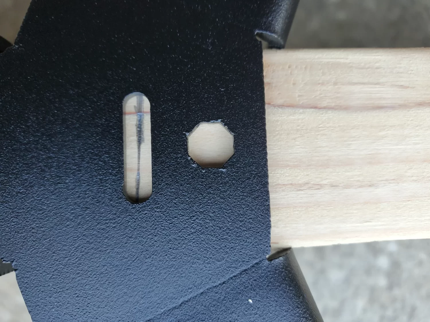 20230510_160846 Close-up of a Heavy Duty Octagon CrossTie Bracket mounted on a light wooden beam. The black metal bracket has a vertical oval hole with a bubble level and an adjacent octagonal hole, set against a concrete background.