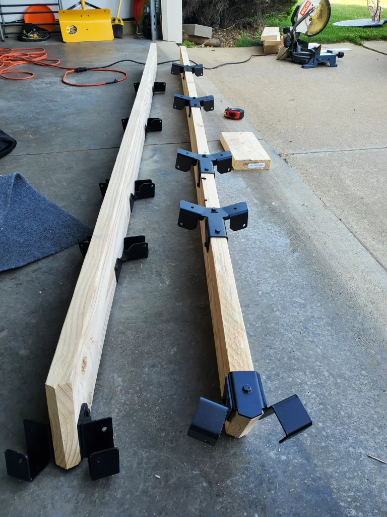 Two long wooden beams on a garage floor, each fitted with multiple Heavy Duty Octagon CrossTie Brackets. In the background near an open garage door are tools, a tape measure, wooden blocks, and cords.
