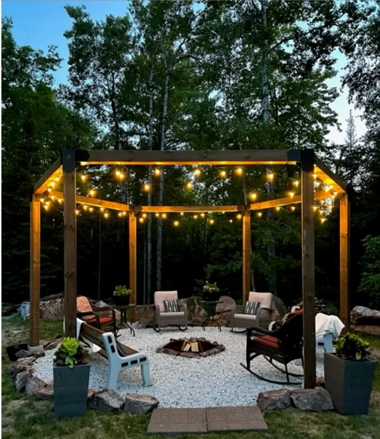 A Surface Mounted, DIY 6x6 Hexagon Gazebo creates a cozy outdoor seating area with string lights and planters on gravel, surrounded by trees and greenery, echoing the ambiance of a firepit space.