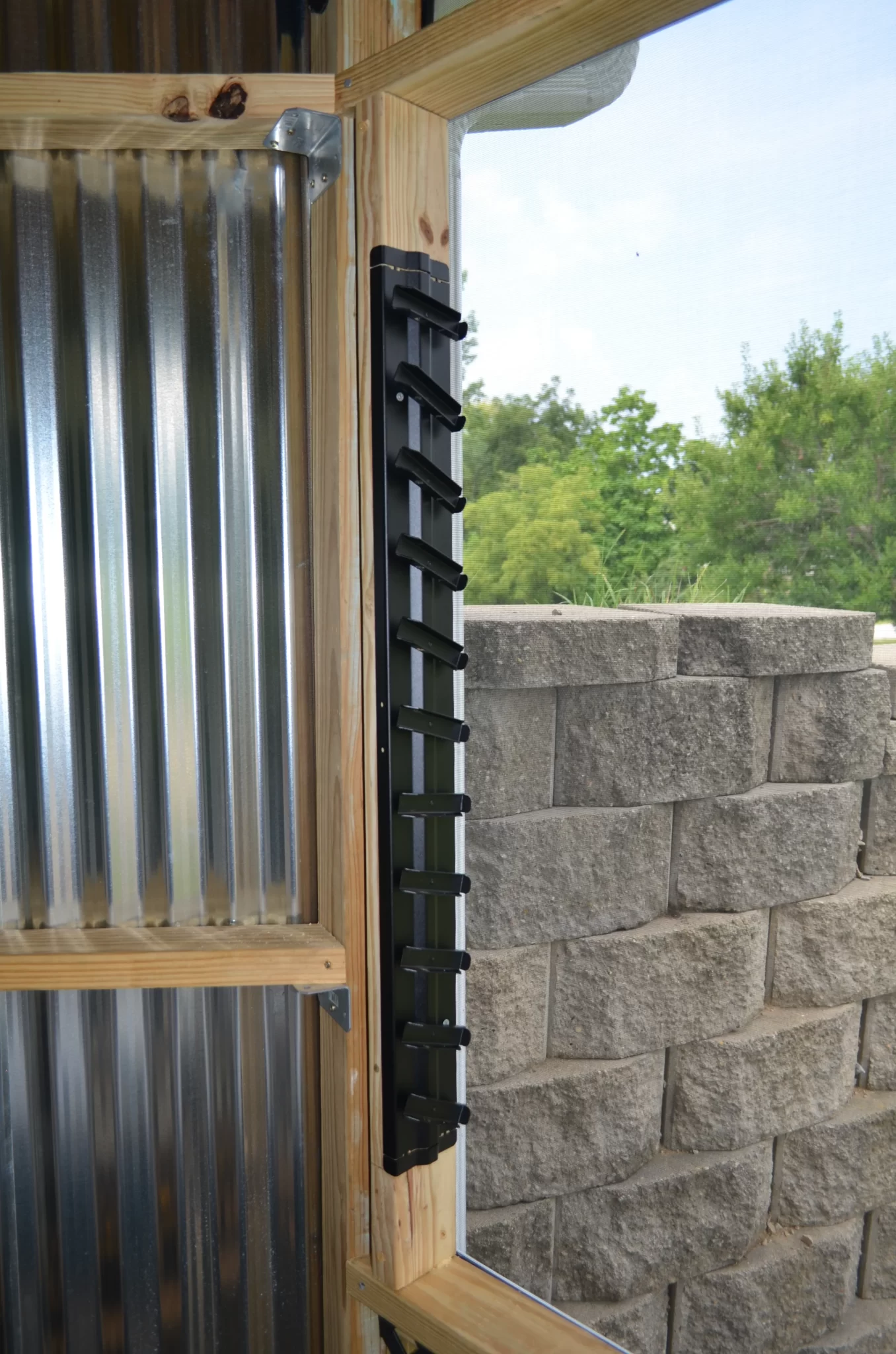 DSC_0070 A black metal ladder on a wooden frame stands by a corrugated metal wall, ideal for reaching your Decorative 36 DIY Louver Kit. A nearby stone retaining wall and vibrant green trees complete the outdoor setting.