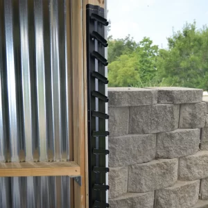 DSC_0070 A black metal ladder on a wooden frame stands by a corrugated metal wall, ideal for reaching your Decorative 36 DIY Louver Kit. A nearby stone retaining wall and vibrant green trees complete the outdoor setting.