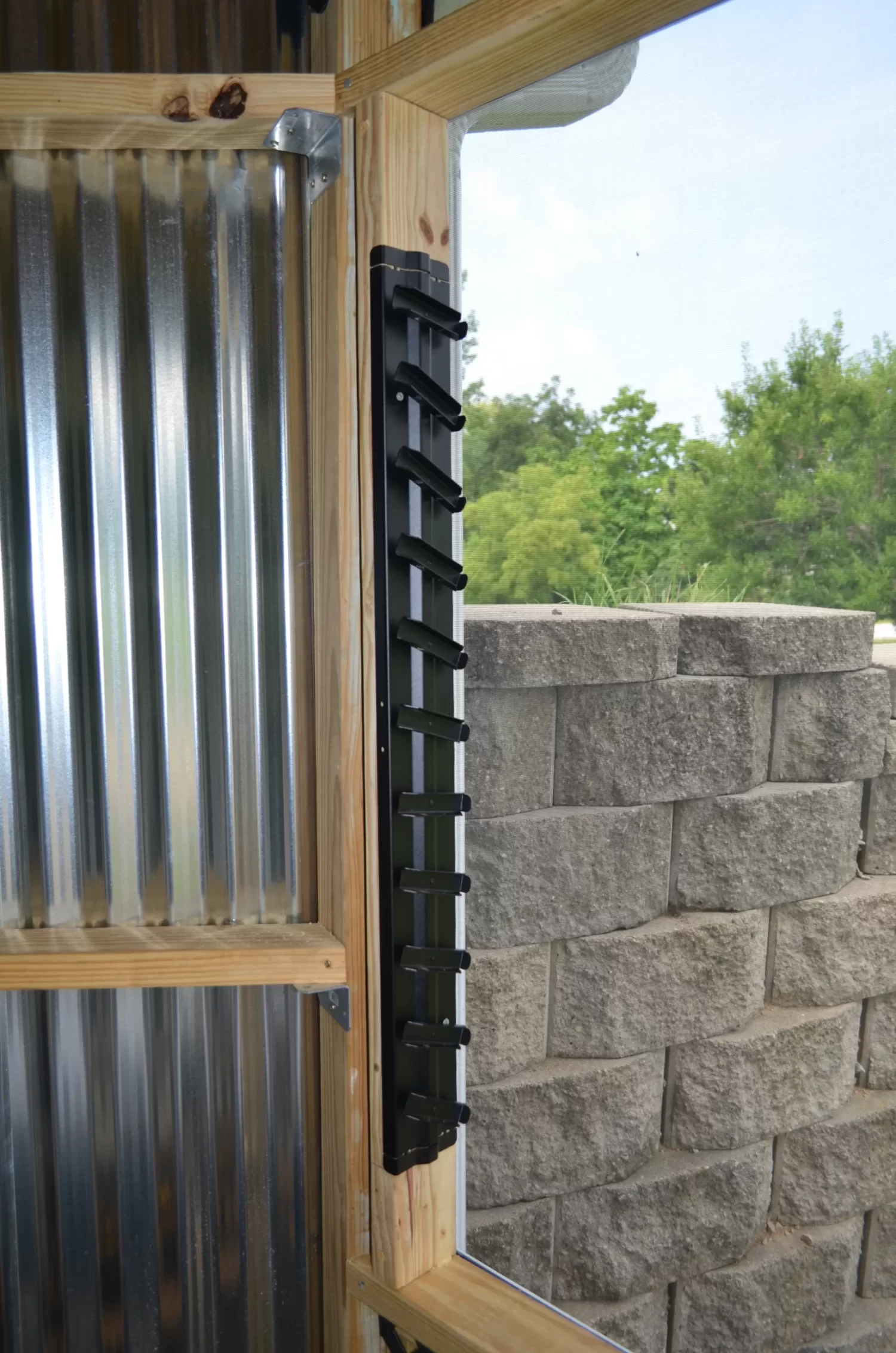 A black metal ladder on a wooden frame stands by a corrugated metal wall, ideal for reaching your Decorative 36 DIY Louver Kit. A nearby stone retaining wall and vibrant green trees complete the outdoor setting.