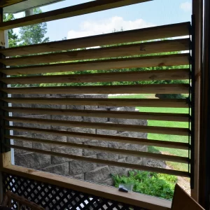 DSC_0086 A horizontal window made with the Decorative 36 DIY Louver Kit features wooden slats and sleek metal and wood framing, offering a partial view of a green lawn, trees, and a stone wall under a blue sky with clouds.