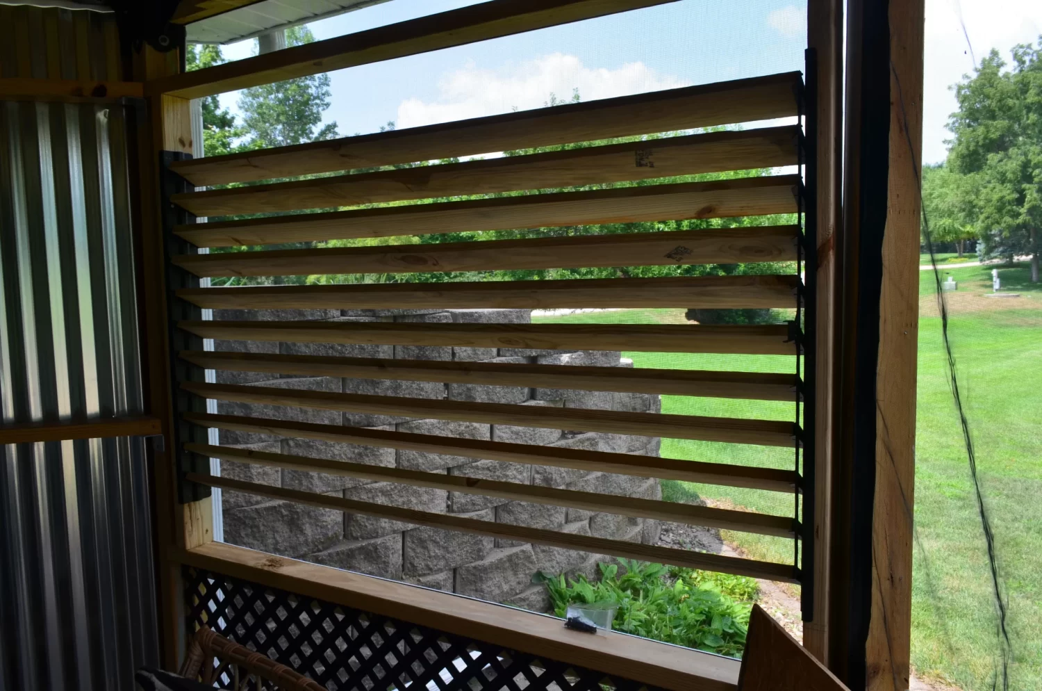 A horizontal window made with the Decorative 36 DIY Louver Kit features wooden slats and sleek metal and wood framing, offering a partial view of a green lawn, trees, and a stone wall under a blue sky with clouds.