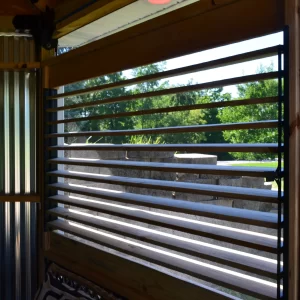 DSC_0104 A sunlit green yard and trees are seen through horizontal black slats of a Decorative 36 DIY Louver Kit, installed in a modern wooden and metal-walled structure, with concrete steps visible outside.