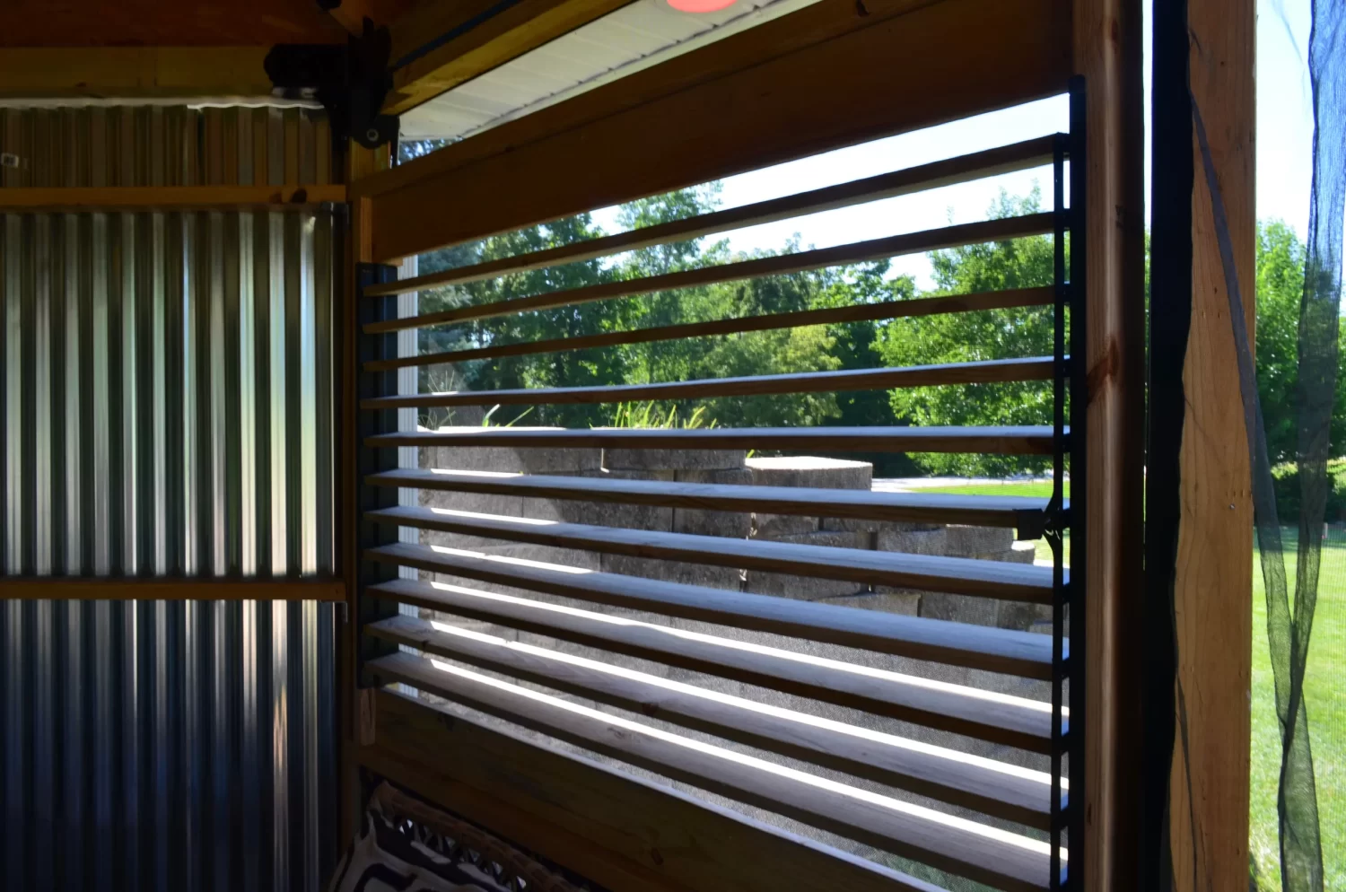 A sunlit green yard and trees are seen through horizontal black slats of a Decorative 36 DIY Louver Kit, installed in a modern wooden and metal-walled structure, with concrete steps visible outside.
