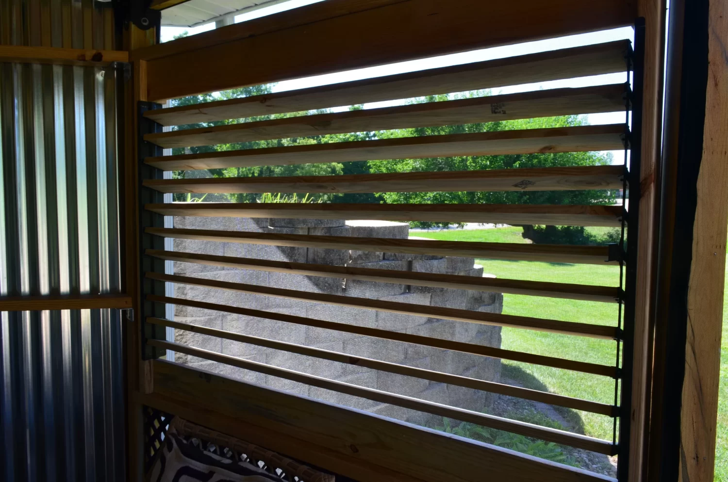 A wooden slatted window with horizontal bars, built with the Decorative 36 DIY Louver Kit, offers a view of green grass, trees, and stacked concrete blocks outside in bright daylight.