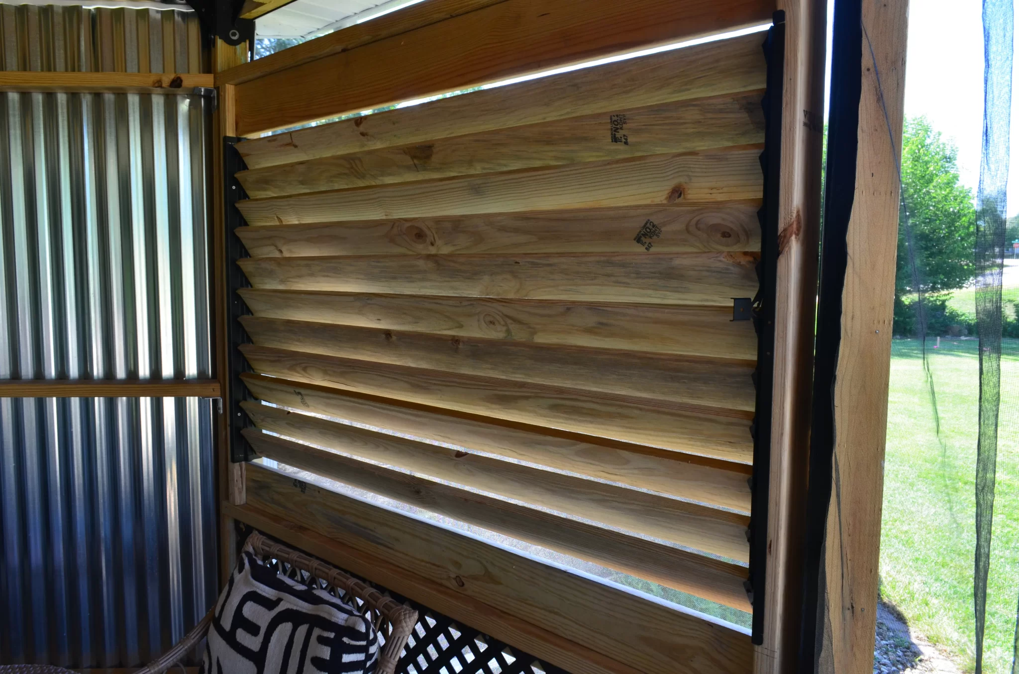 DSC_0114 A close-up of a wooden louvered window, built with the Decorative 36 DIY Louver Kit, installed in a partially enclosed patio with corrugated metal walls and outdoor greenery visible through the slats.