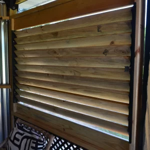 DSC_0114 A close-up of a wooden louvered window, built with the Decorative 36 DIY Louver Kit, installed in a partially enclosed patio with corrugated metal walls and outdoor greenery visible through the slats.