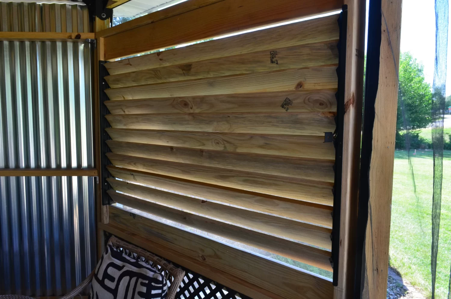 A close-up of a wooden louvered window, built with the Decorative 36 DIY Louver Kit, installed in a partially enclosed patio with corrugated metal walls and outdoor greenery visible through the slats.