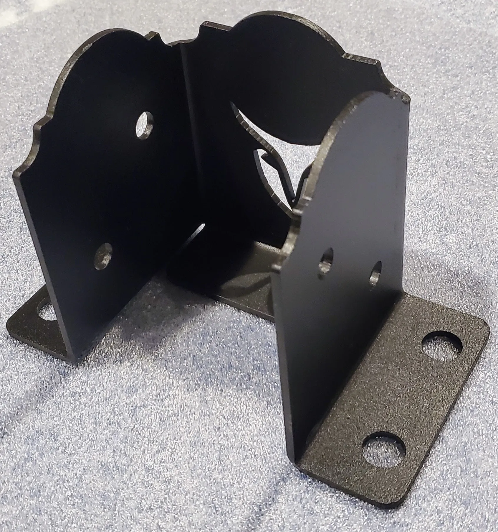 4LC-2 (1) The Heavy Duty 4x4 Post Anchor is a black metal bracket with decorative edges, four mounting holes on its base and vertical sides, shown on a textured grey surface.