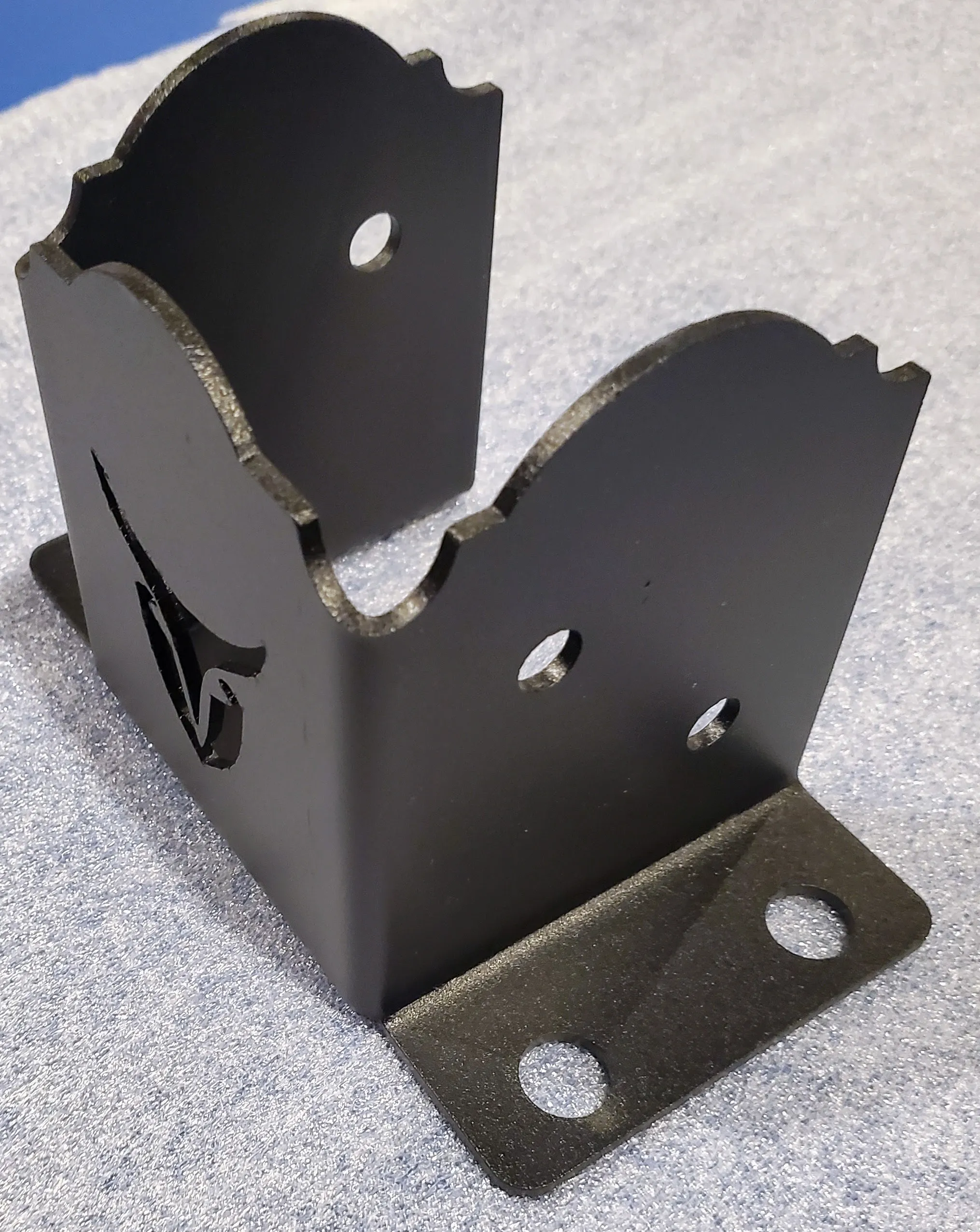4LC-3 (1) The Heavy Duty 4x4 Post Anchor is a black metal bracket with curved edges, multiple circular holes, a side cutout design, and a flat base with two mounting holes, shown on a textured grey surface.