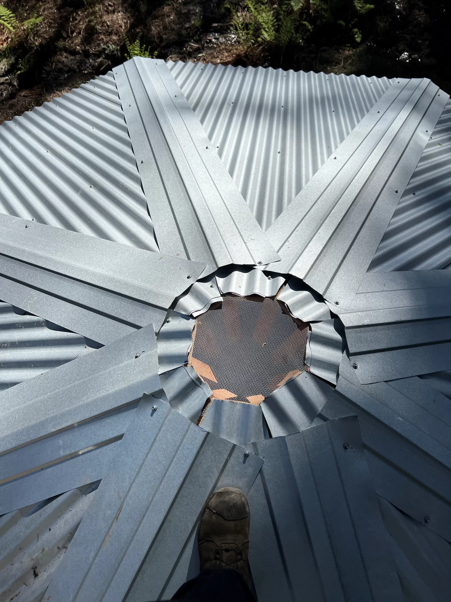 A person’s boot stands on the tank roof4, a corrugated metal structure with a star-shaped design and open circular center, allowing mesh and ground to be seen below as sunlight highlights its metallic surface.