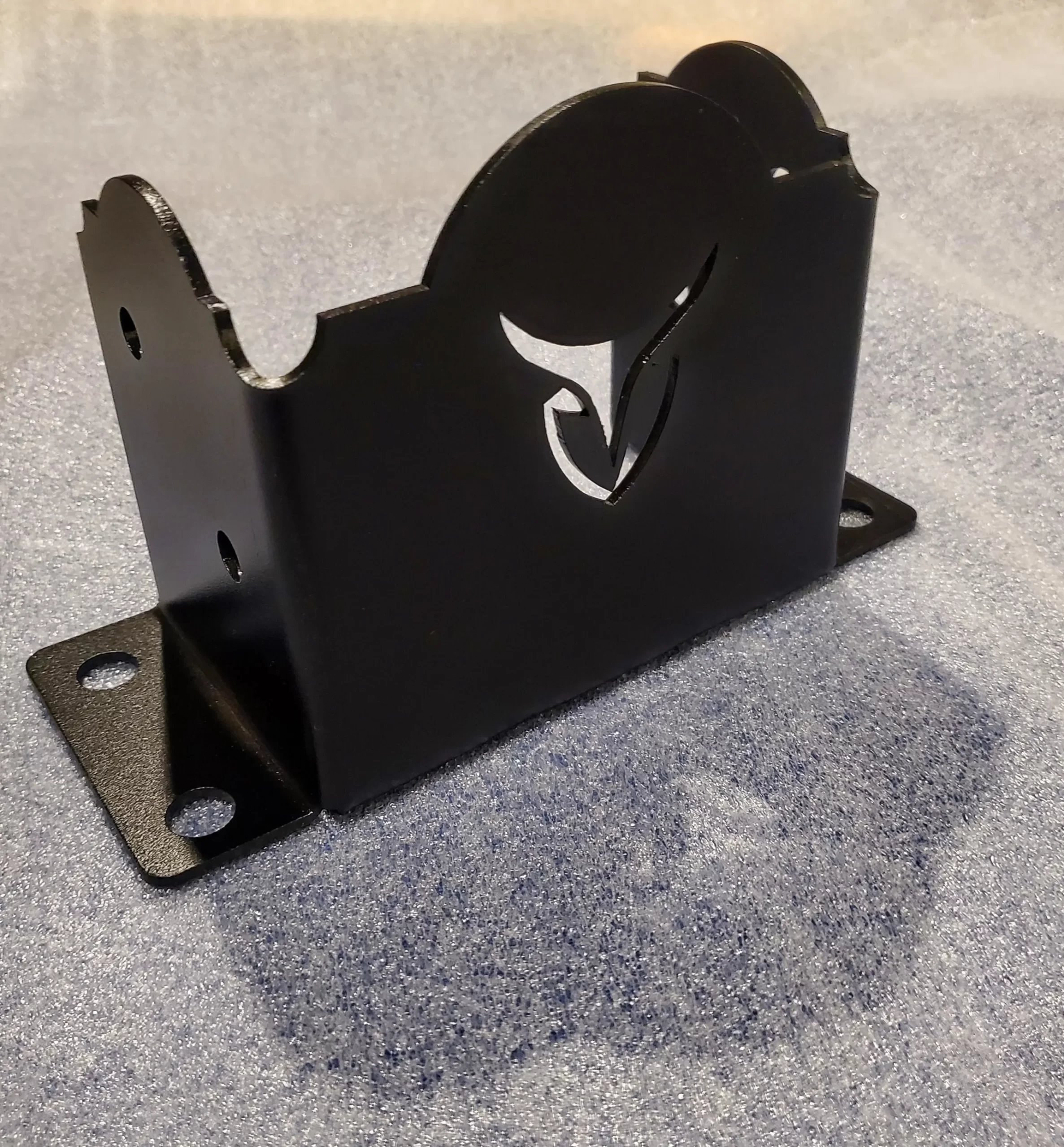 20251117_181038 A black metal bracket with decorative curved edges and a cutout floral design stands on a speckled surface. The bracket has a flat base with four mounting holes for installation.