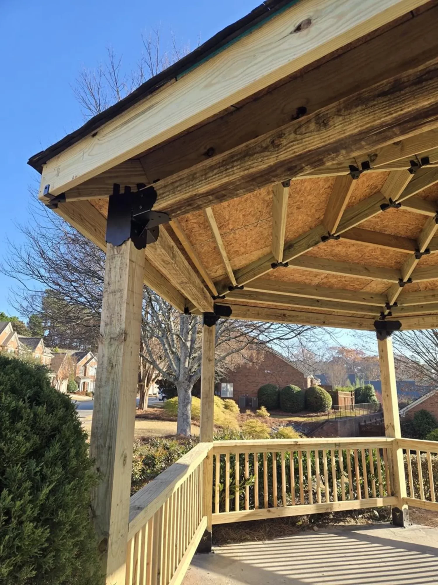 A wooden gazebo with a partially finished roof and exposed beams stands near a bush, with houses and trees visible in the background under a clear blue sky.