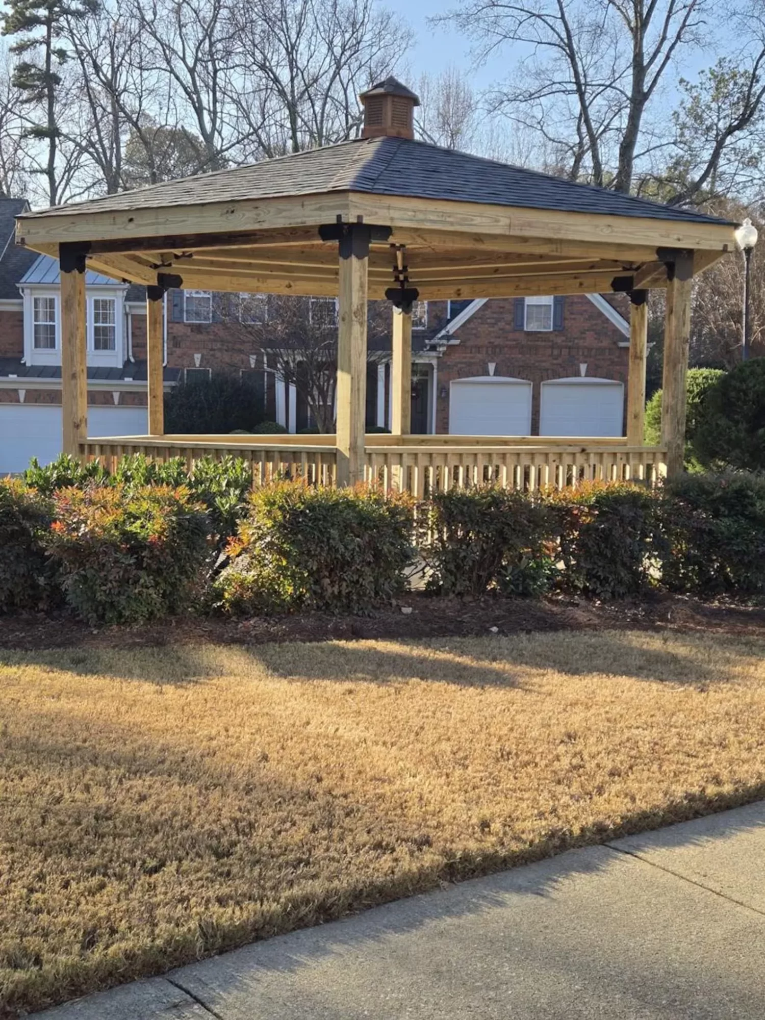 A wooden gazebo with a dark shingled roof stands in front of a row of brick garages. Shrubs and a patch of grass surround the gazebo, and tall trees without leaves are visible in the background.