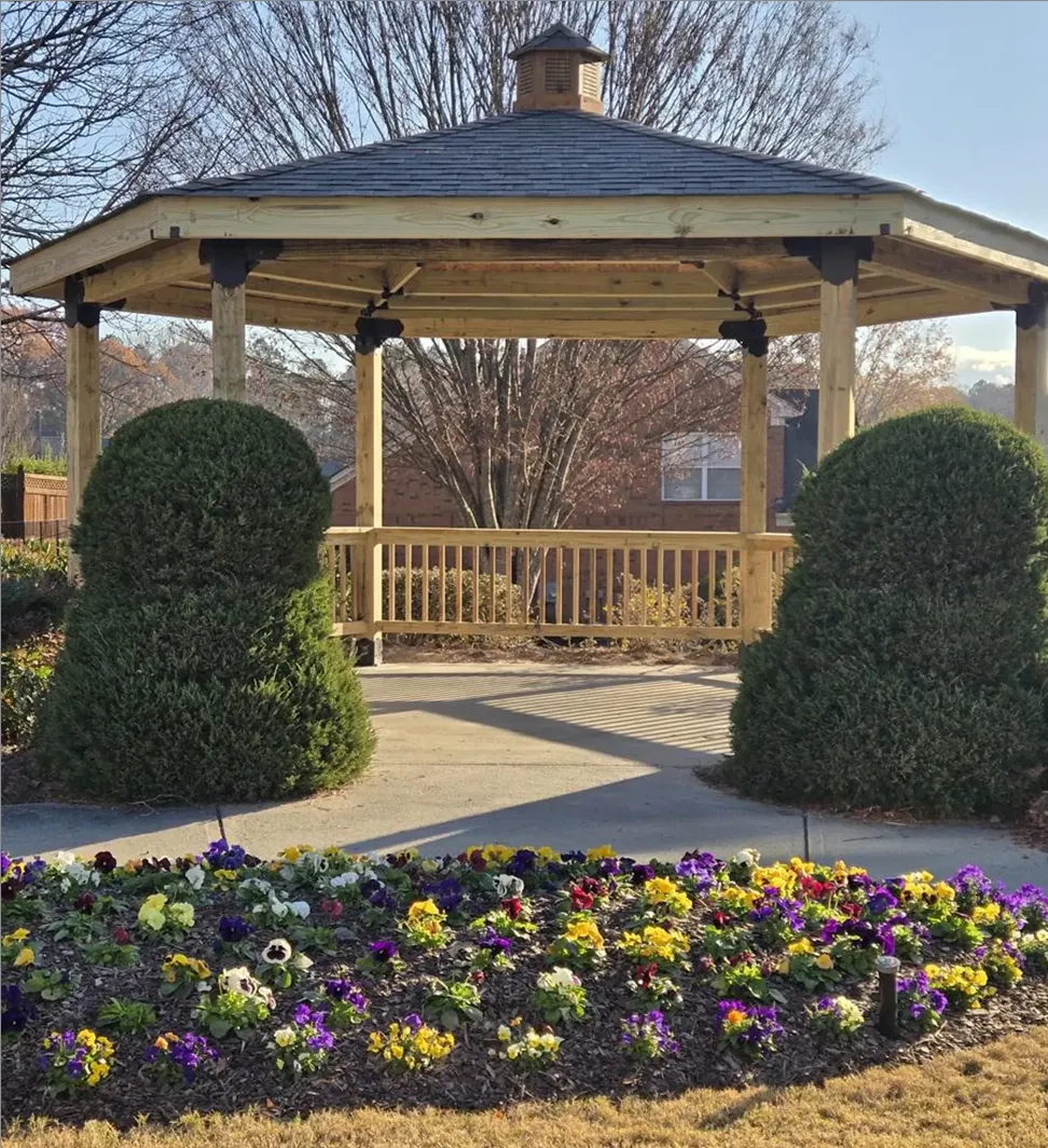 A wooden gazebo with a shingled roof stands behind two large, neatly trimmed bushes and a vibrant flowerbed filled with yellow, purple, and white flowers on a sunny day.
