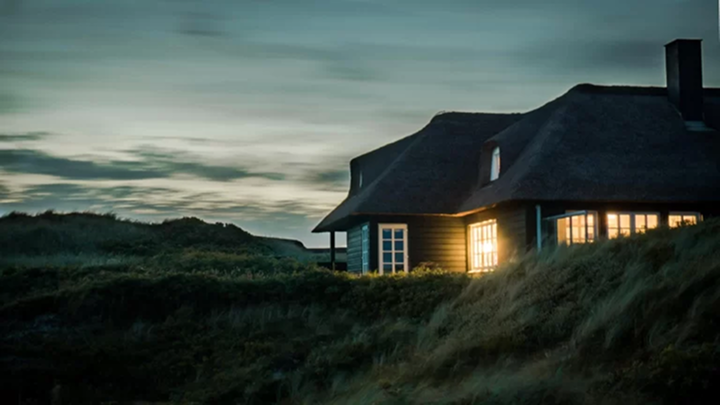 A cozy house with a thatched roof glows warmly at dusk, nestled among grassy dunes under a cloudy evening sky.