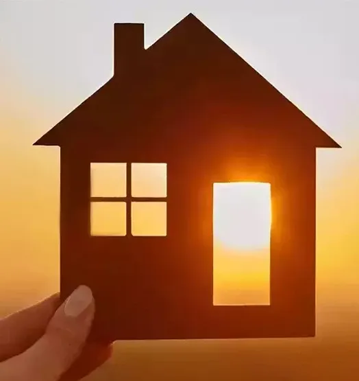 A hand holds a cutout silhouette of a house against a bright sunset sky, with sunlight shining through the windows and door openings.